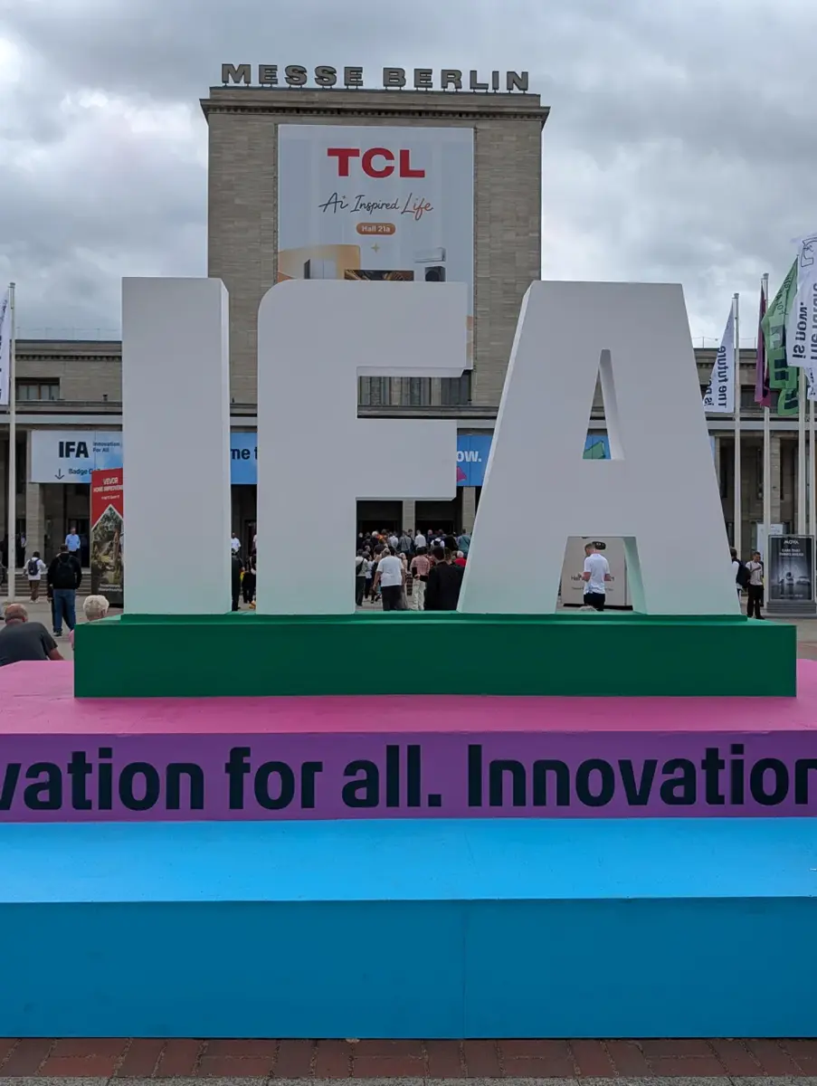 Die großen IFA-Buchstaben auf einem grün-rosa Podest vor dem Eingang Nord, davor Besucher, im Hintergrund das Gebäude mit TCL-Werbung, bewölkter Himmel.