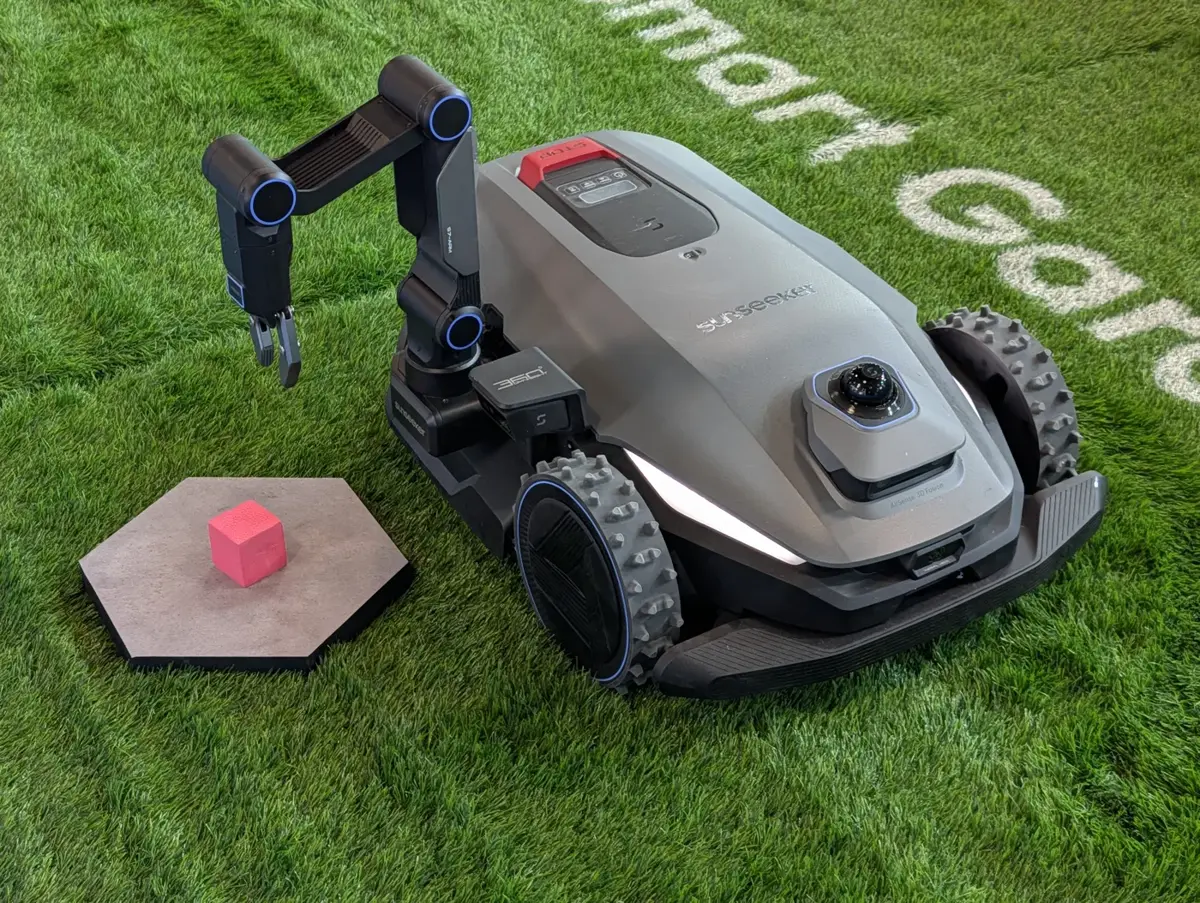 Sunseeker-Mähroboter auf Kunstrasen mit einem schwarzen Roboterarm auf dem Heck, dessen Greifer über einem roten Würfel auf einer sechseckigen Platte schwebt, seitlich ein Modul mit der Aufschrift 360.