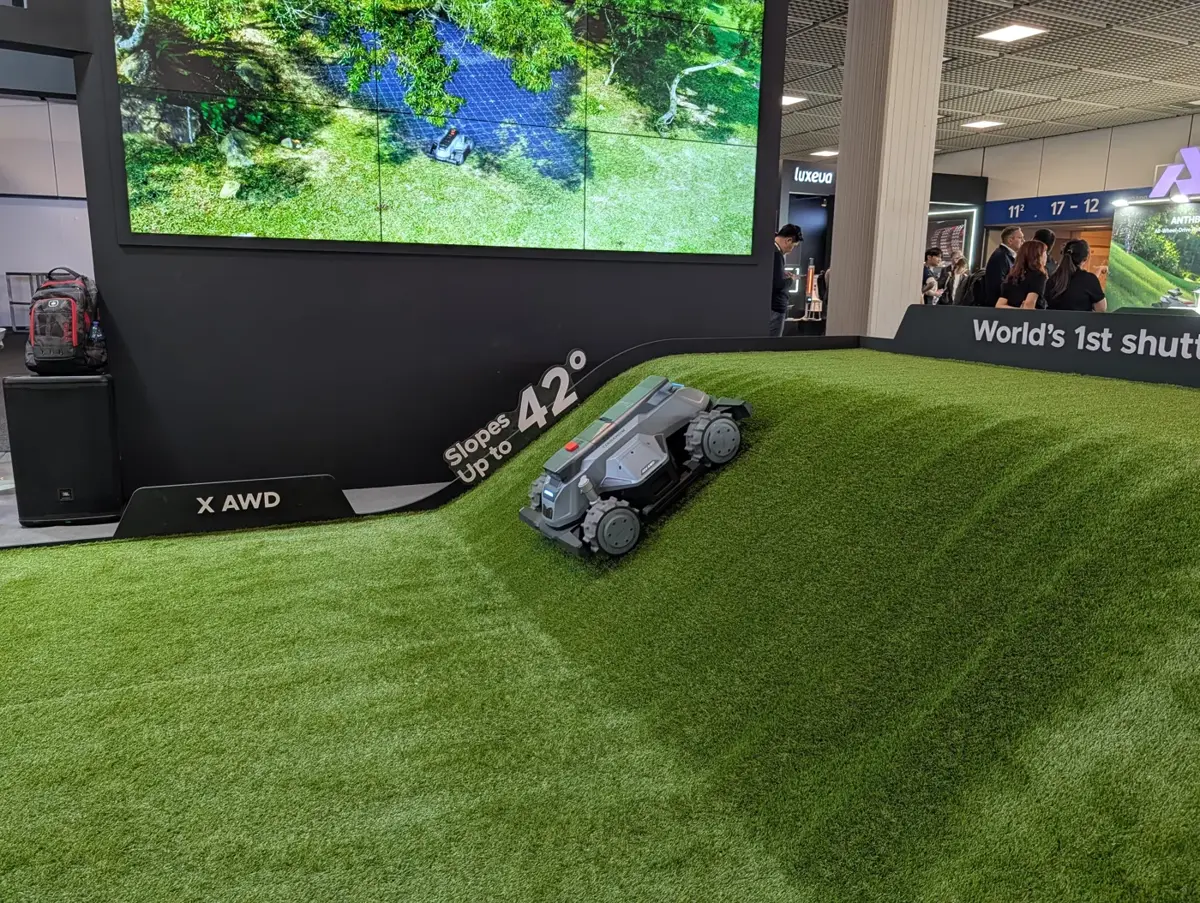 TerraMow X AWD, ein graugrüner Allrad-Mähroboter mit rotem Stoppknopf, steht schräg auf einer steilen Kunstrasenrampe, daneben das Schild Slopes up to 42 Grad und die Beschriftung X AWD, dahinter ein Bildschirm mit Gartenkarte.